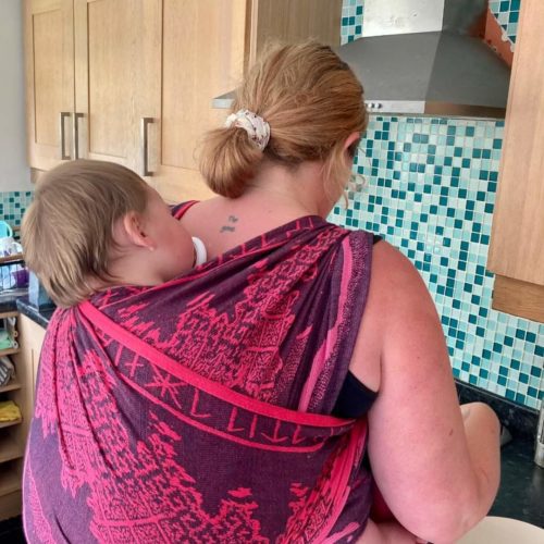 woman cooks with baby wrapped on her back in pink and navy woven sling
