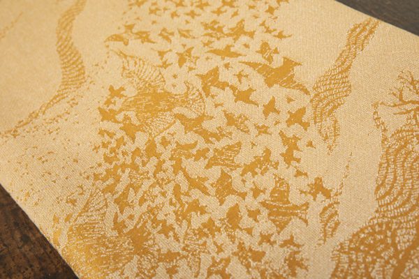 A woven wrap that has a shimmery pale grey linen and merino blend weft against a mustard-gold organic cotton warp. Our Murmuration design shows a starling murmuration over a wintery treeline
