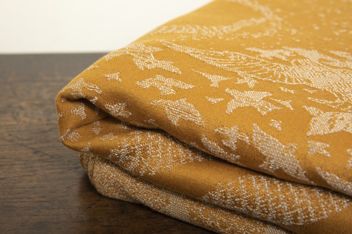 A woven wrap that has a shimmery pale grey linen and merino blend weft against a mustard-gold organic cotton warp. Our Murmuration design shows a starling murmuration over a wintery treeline