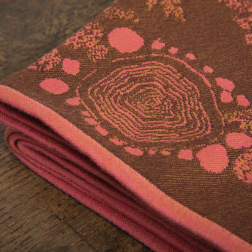 A woven wrap with a warm salmon pink and dark brown design of a rainy hillside with fossils and fractals underground and pylons above