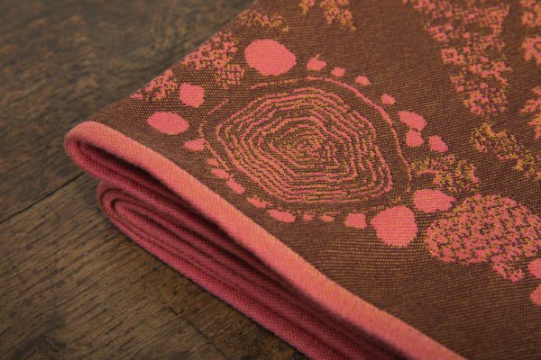 A woven wrap with a warm salmon pink and dark brown design of a rainy hillside with fossils and fractals underground and pylons above