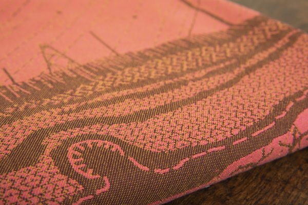 A woven wrap with a warm salmon pink and dark brown design of a rainy hillside with fossils and fractals underground and pylons above