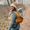 mustard gold and brown woven wrap worn in a double hammock back carry by a smiling white woman with brown hair and woolly hat